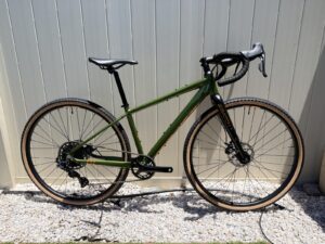 The Batch GB.1 is a $999 Gravel Bike Built to Get You Riding Batch GB.1 gravel bike shown from the non-drive side, olive-green aluminum frame with copper accents and tan-wall gravel tires, leaning against a light-panelled wall.