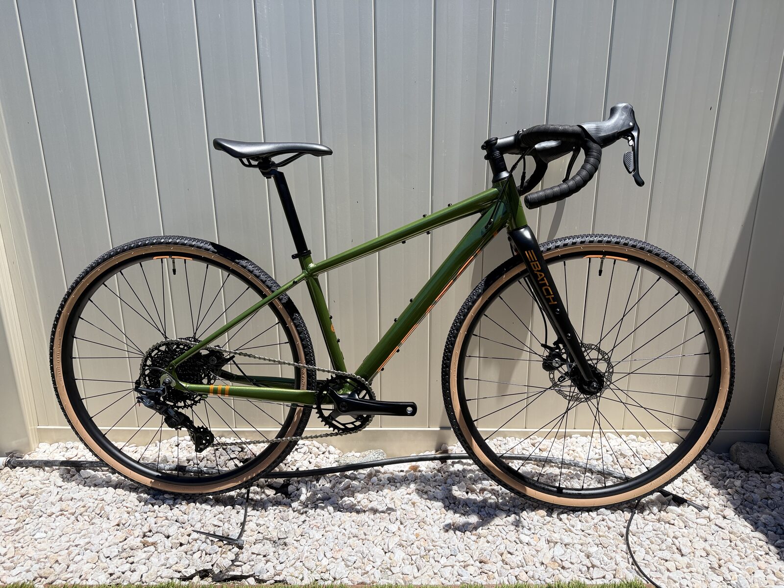 Batch GB.1 gravel bike shown from the non-drive side, olive-green aluminum frame with copper accents and tan-wall gravel tires, leaning against a light-panelled wall.