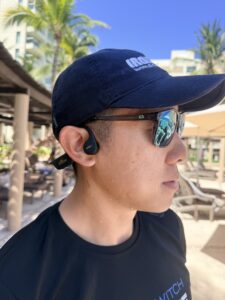 Reviewer in an IRONMAN cap and Slowtwitch shirt wearing the H2O Audio TRI 2 PRO open-ear bone conduction headphones on a resort pool deck.